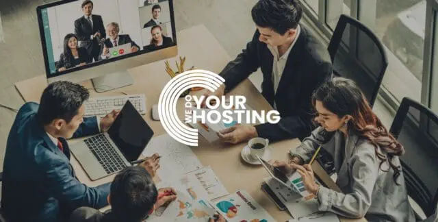 WeDoYourHosting white logo on background with workers sat at a desk together with paper graphs on desk. Mac monitor showing a video call with other workers on screen