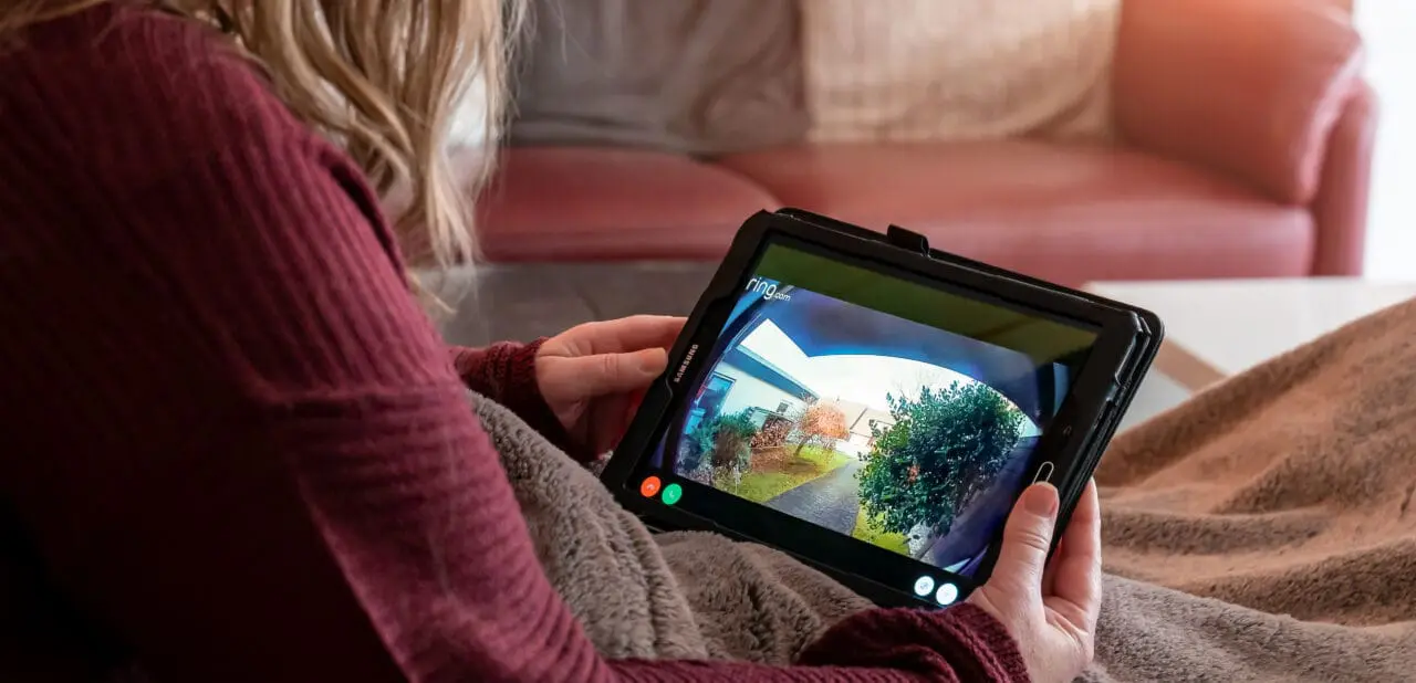 A woman is lying on the couch. She looks on tablet. Display shows shot of the surveillance camera ring from Amazon.