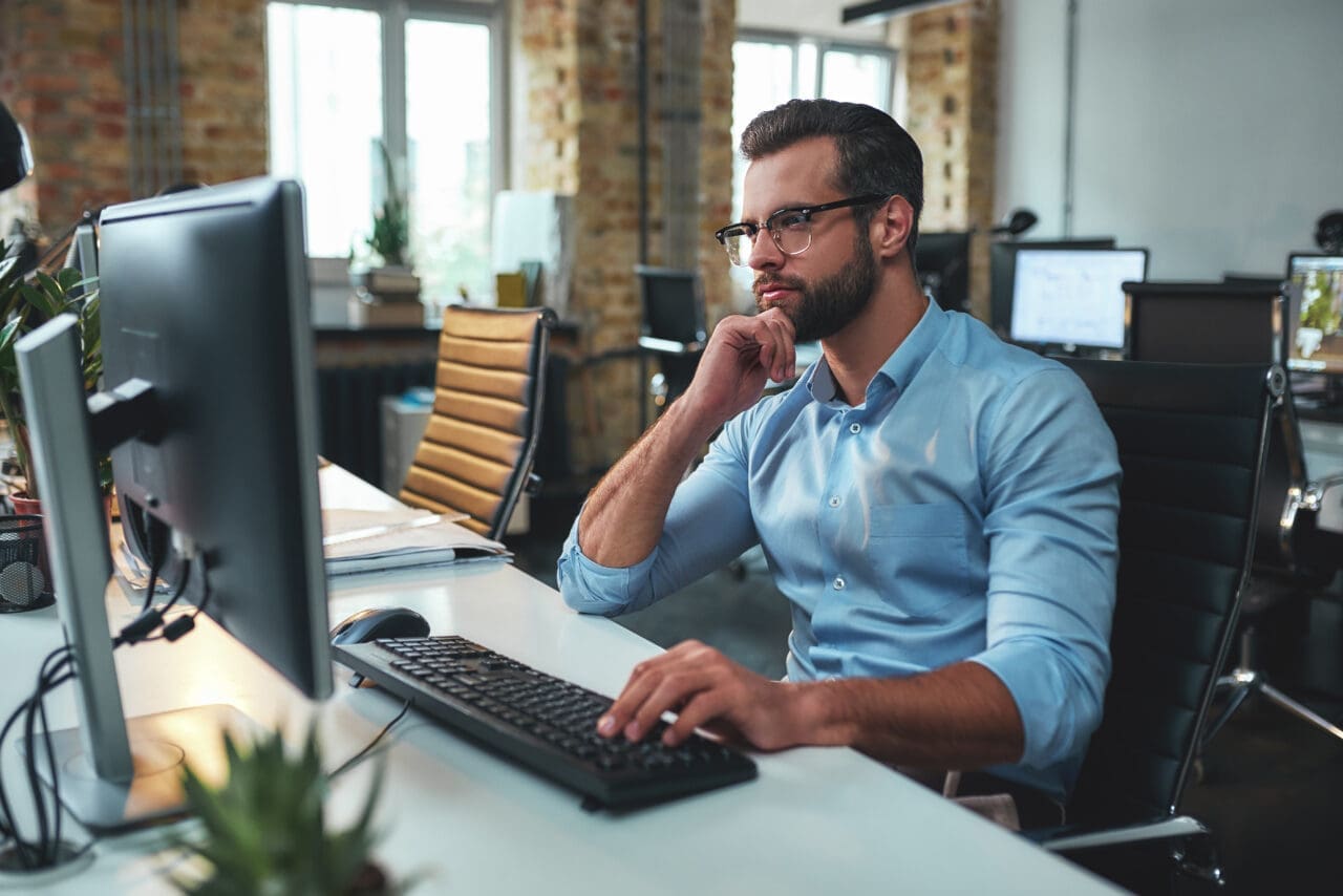 New project. Side view of young bearded man in eyeglasses and formal wear working on computer and touching his chin while sitting in the modern office. Business concept. Work concept. Modern technologies