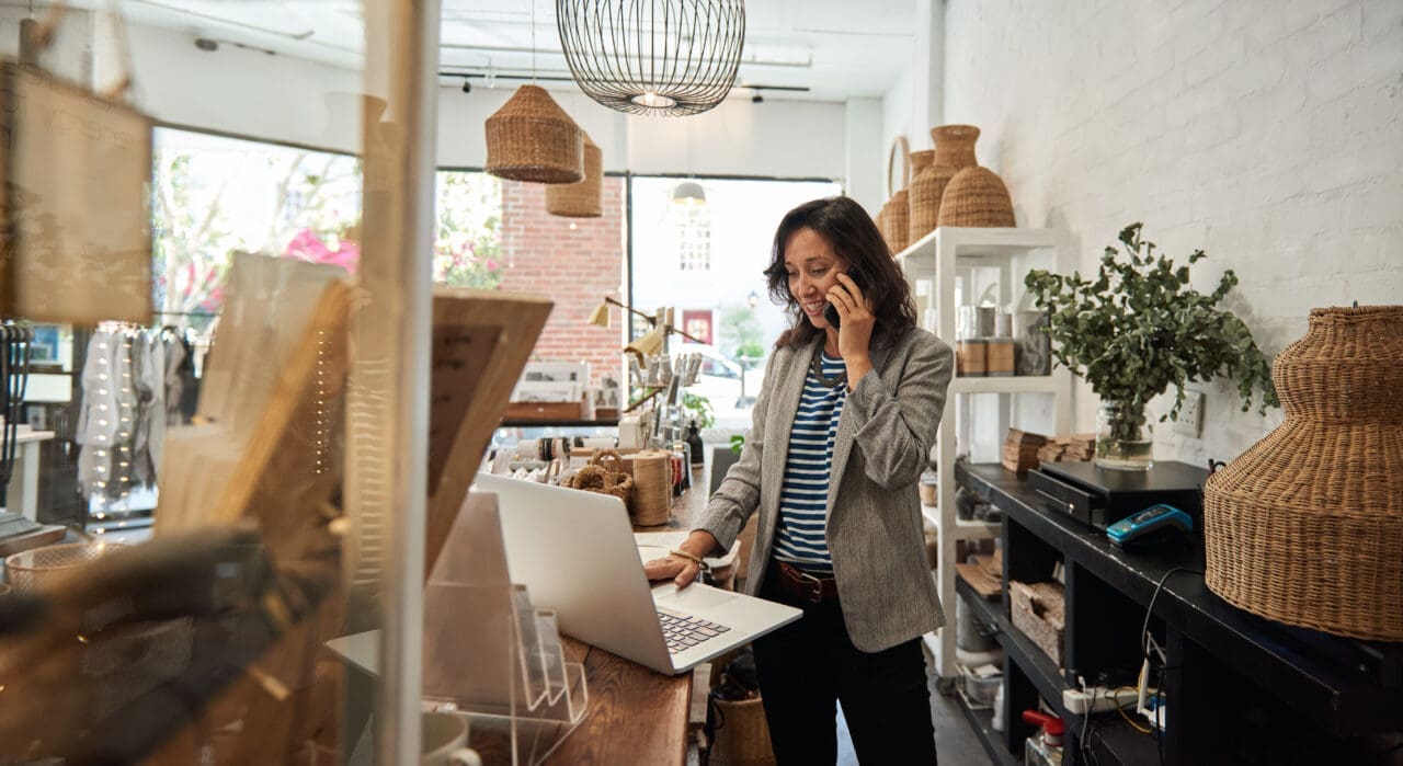 Smiling Asian woman talking on a cellphone in her store, whilst working on her Mac laptop