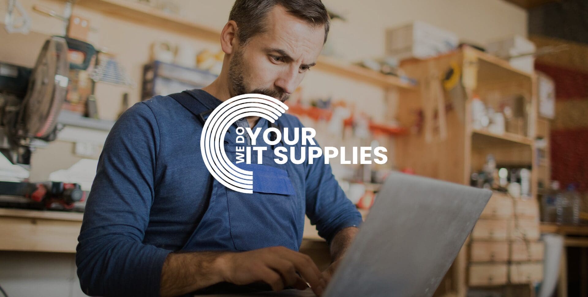 WeDoYourITSupplies white logo on background of man in workshop, typing on his laptop