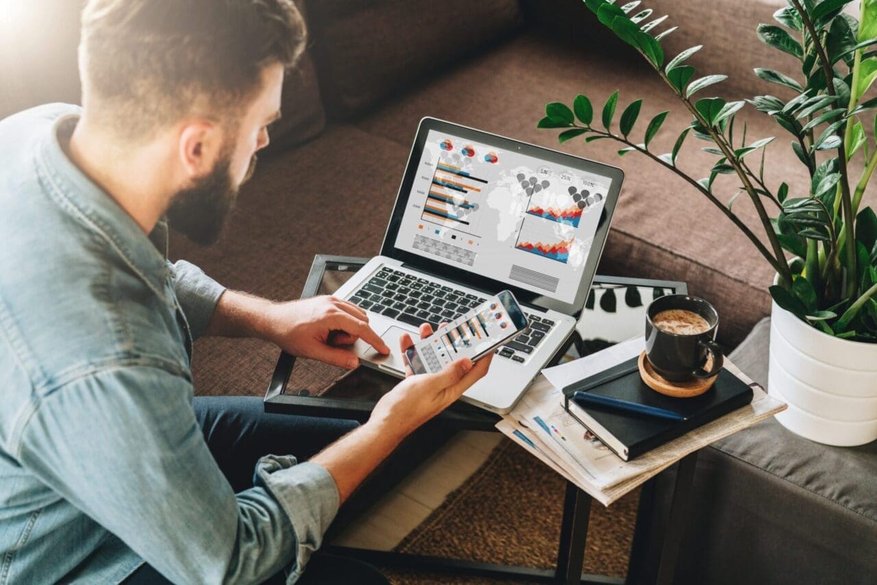 Man, entrepreneur, freelancer sits at home on couch at coffee table, uses smartphone, working on laptop with graphs