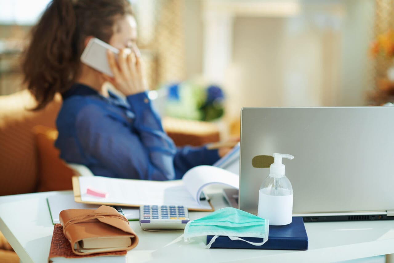 Blurred background of woman sat at home, talking on her mobile phone. Front of the image is a laptop on a desk, with hand sanitiser and a face mask