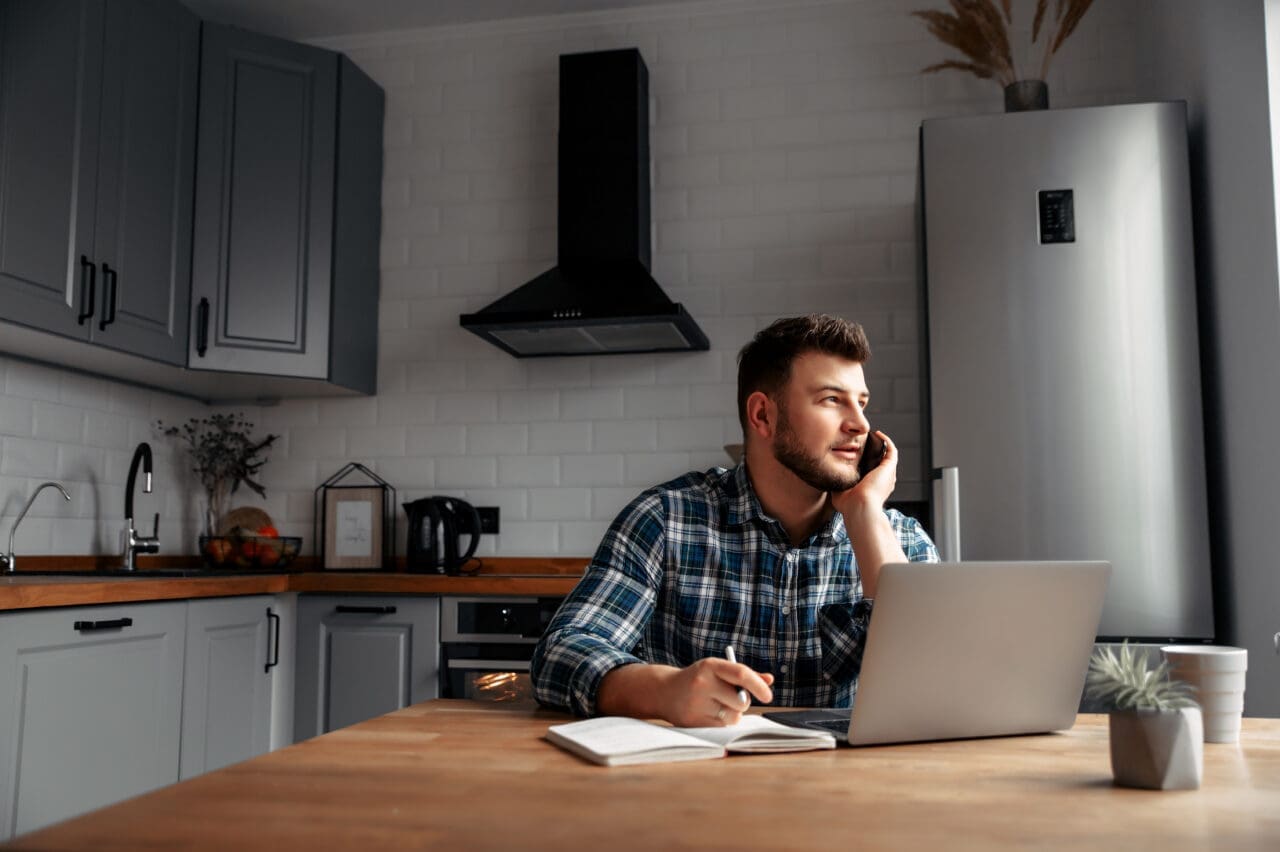 Young male sat at kitchen table, with mobile phone to his ear. Mac laptop is placed on the table, with an open notebook placed next to it