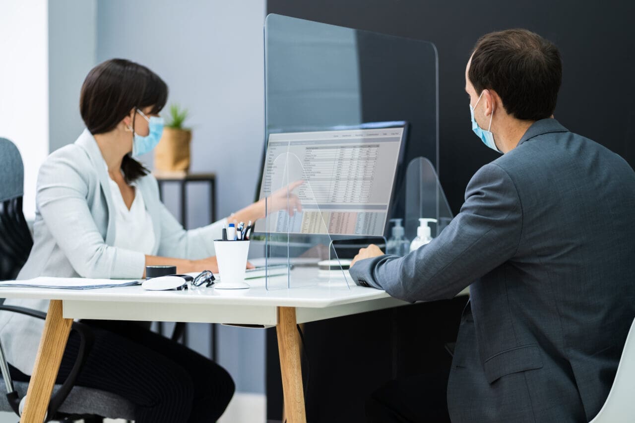 Financial Advisor Meeting With Client Customer. Both wearing face masks and plastic screen in-between them placed on desk