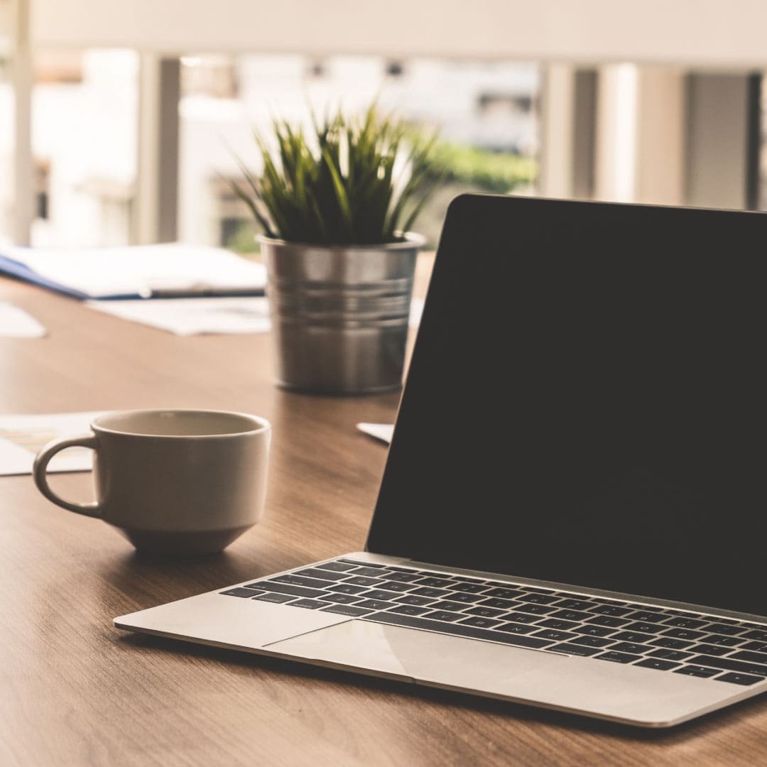 Mac laptop on brown desk with coffee cup and plant besides it