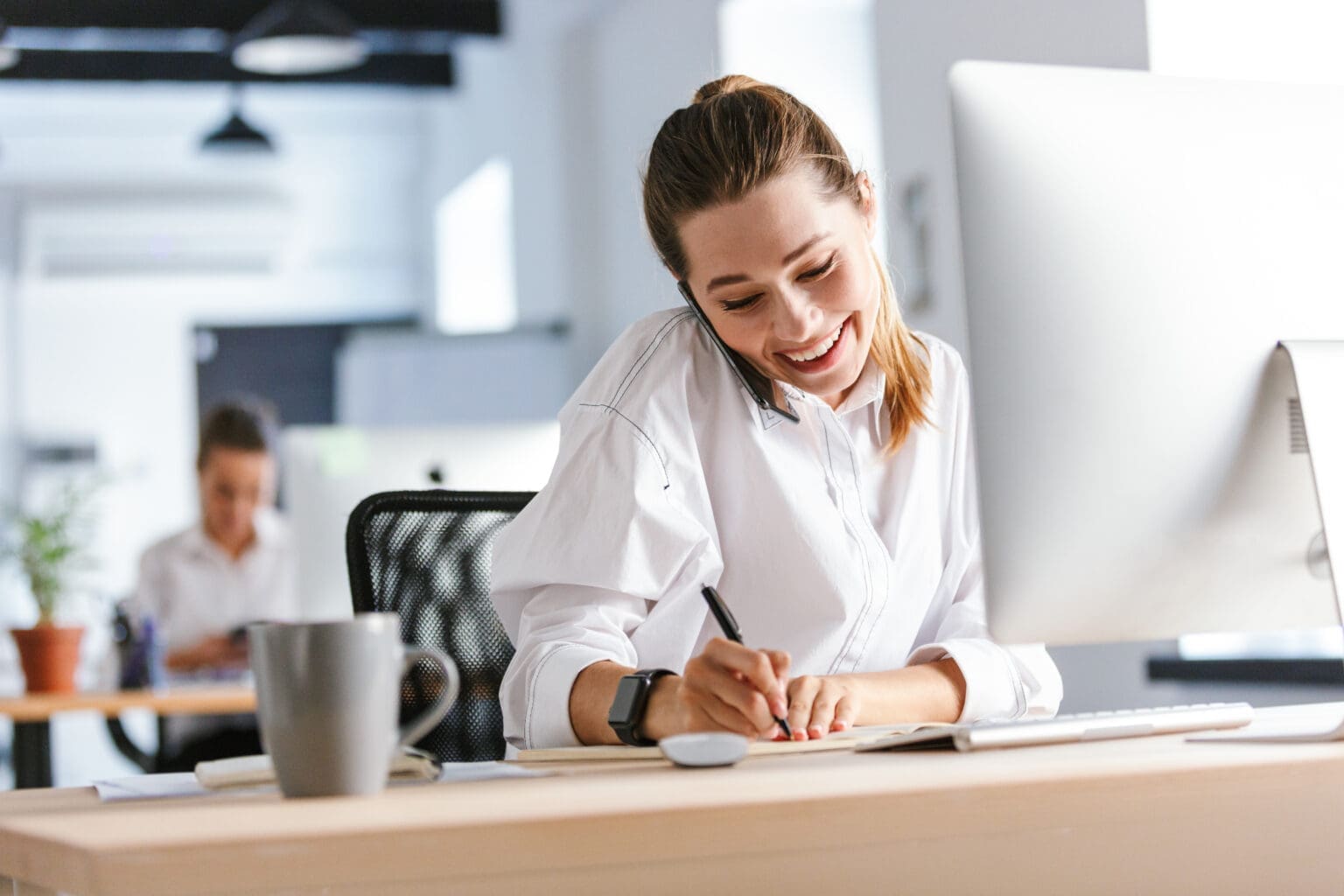 Cheerful young businesswoman sitting at her workplace with Mac computer, talking on phone, taking notes