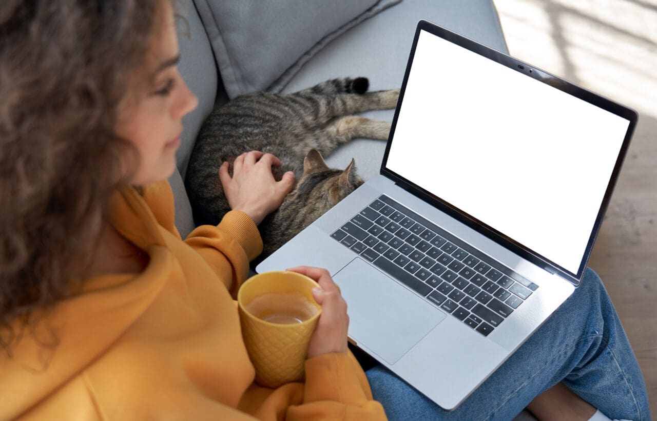 Young girl relaxing on sofa with cat looking at Mac laptop screen.