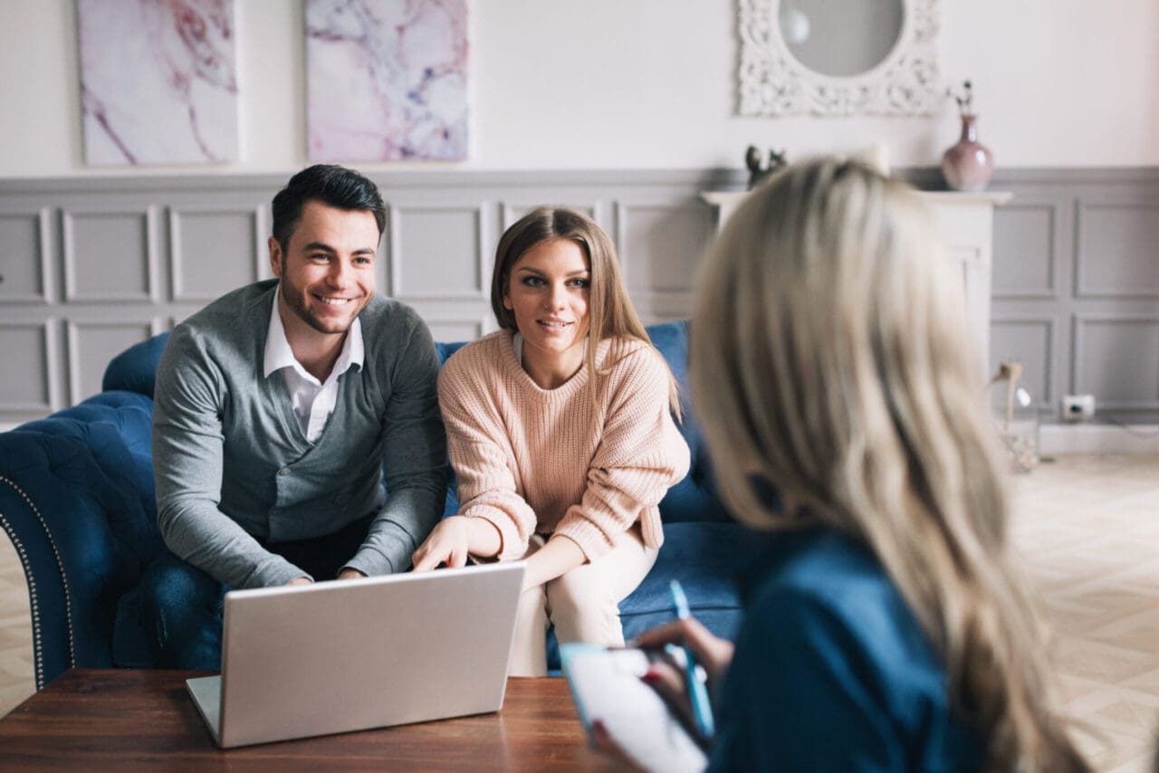 Image of young couple sat on sofa smiling, with laptop on coffee table. Business woman sat opposite them