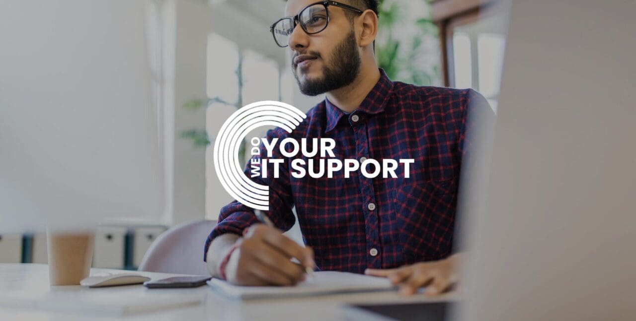 WeDoYourITSupport white logo on background of man writing on notebook, with Mac laptop in front of him