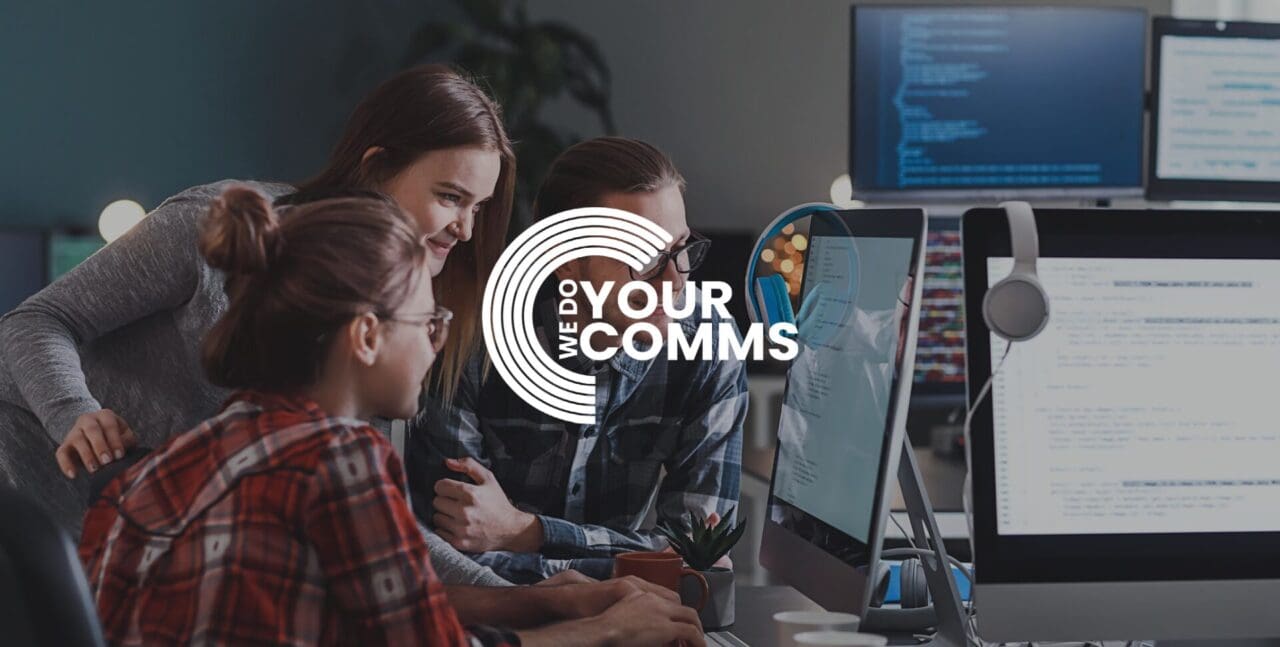 WeDoYourComms white logo on background with young workers smiling and working together on Mac computer