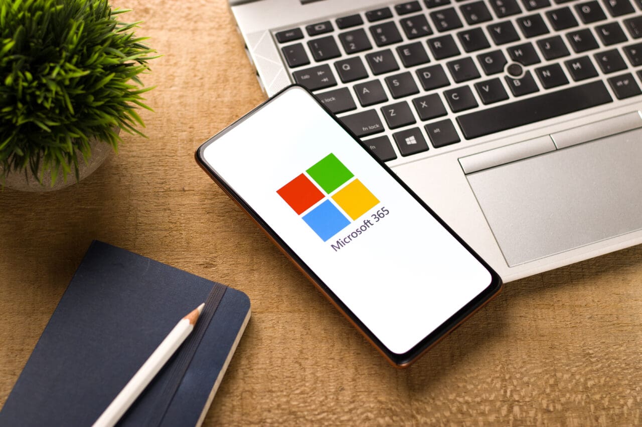 Microsoft 365 logo on screen of mobile phone, placed on top of laptop.