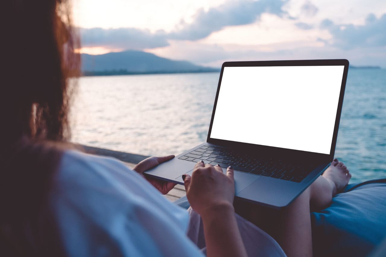 Image of a woman using laptop with blank white desktop, sat in front of calm water