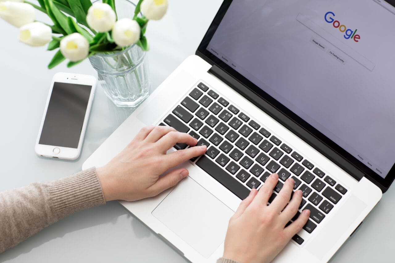 Woman typing on a Mac laptop, with Google search engine on the screen. Mobile phone next to it with some flowers