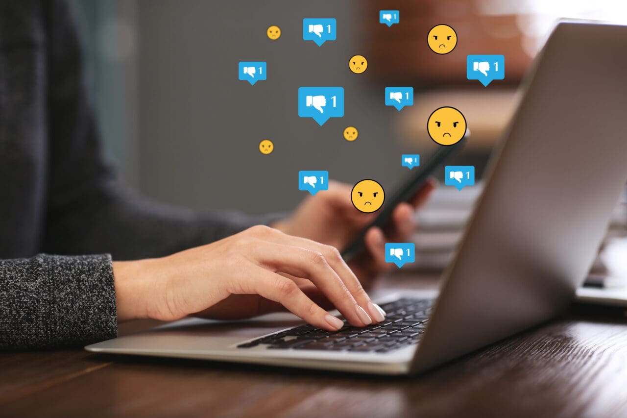 Social media dislike reaction. Woman using laptop and mobile phone at table, closeup. Thumbs down and angry face emoji illustrations over device