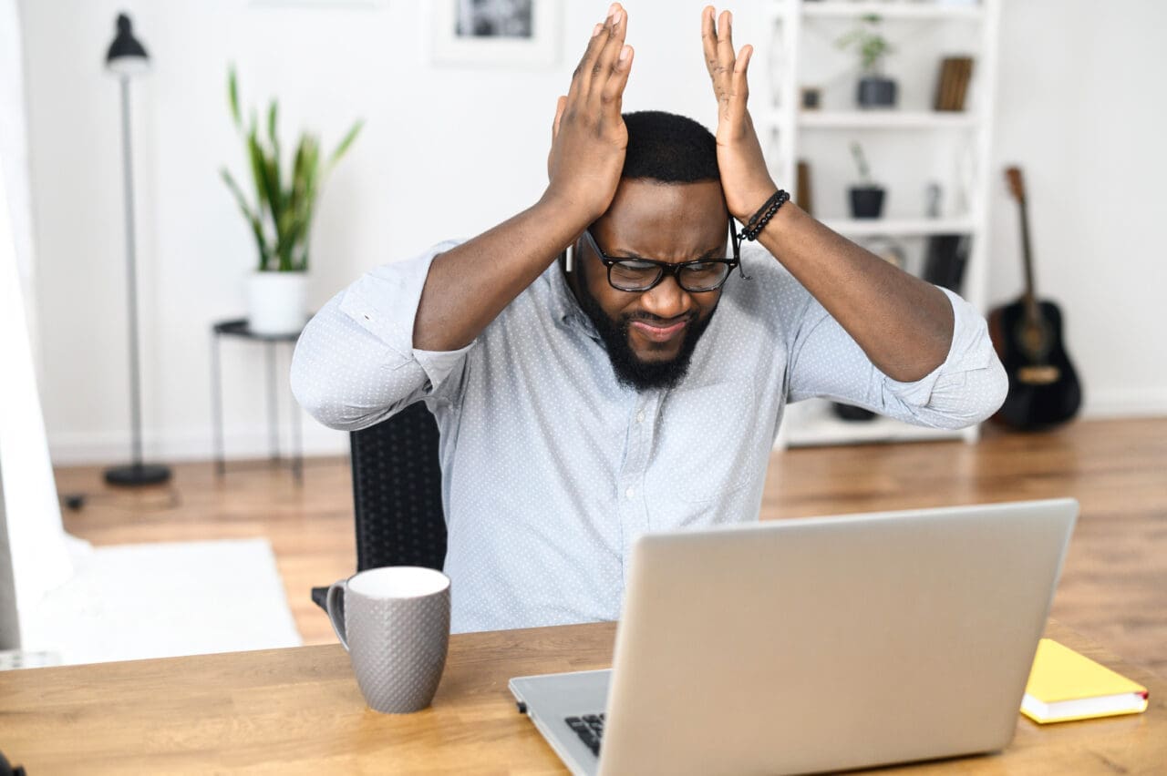 Frustrated businessman in smart casual holding head in hands, sitting alone at the modern office desk with an open laptop, stressing out from occurred computer issues