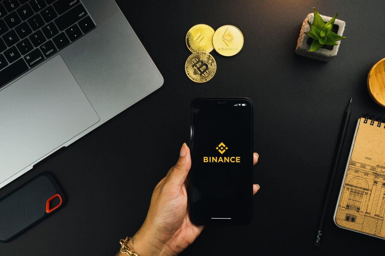 Girl holding a smartphone with Binance cryptocurrency exchange app on the screen on black table with a cryptocurrencies coins and computer. Office environment.