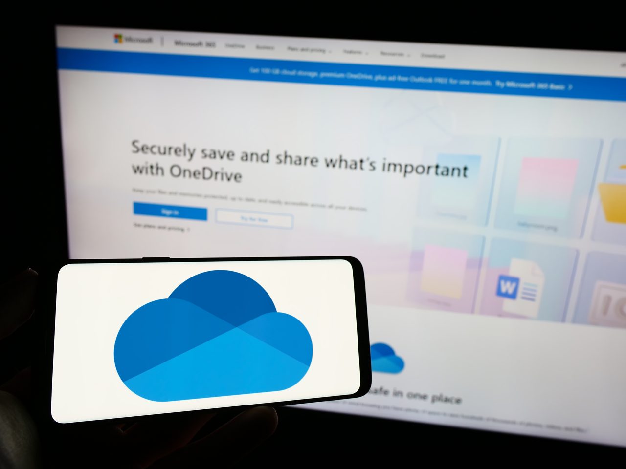 Person holding cellphone with logo of file hosting service Microsoft OneDrive on screen in front of business webpage. Focus on phone display.