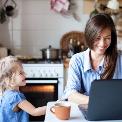 Image of young woman sat at kitchen table, working on her laptop. Smiling and laughing with her young daughter next to her